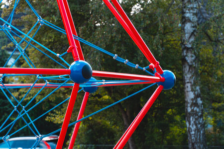 Spielplatzspielgerät in Rot-Blau Spielgerät im Schulhof mit roten und blauen Stangen und Verbindungsseilen, umgeben von Bäumen und Grünanlagen.