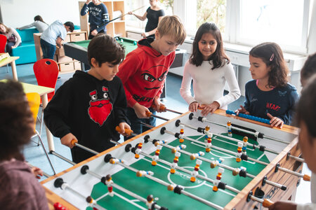 Kinder beim Tischfußballspielen Kinder spielen an einem Tischfußballspiel in einem hellen Raum der Schule. Im Hintergrund sind weitere Schüler und Aktivitäten zu erkennen.