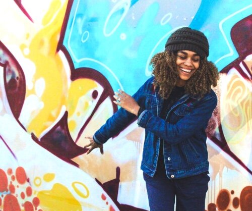 Frau vor Graffiti-Wand Lächelnde Frau in einer Jeansjacke und Mütze, die vor einer bunten Graffiti-Wand steht und eine Hand ausstreckt.
