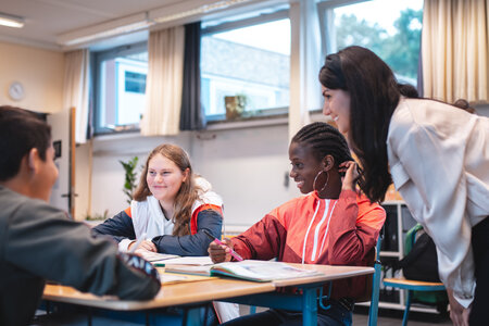 Schüler im Klassenzimmer Schülerinnen und Schüler arbeiten zusammen an Schreibtischen in einem Klassenraum. Eine Lehrerin steht bei ihnen und unterstützt die Gruppe.