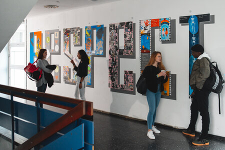 Kunstinstallation in der Schule Schülerinnen und Schüler stehen in einem Flur vor einer Wand mit Kunstwerken. Einige unterhalten sich, während andere die Bilder betrachten.